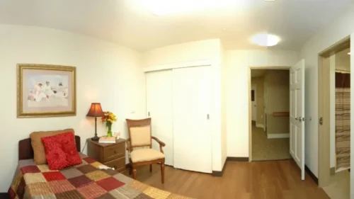 A panoramic view of a bedroom with a plaid bedspread, a chair, a sliding closet door, and an open doorway leading to an adjacent room